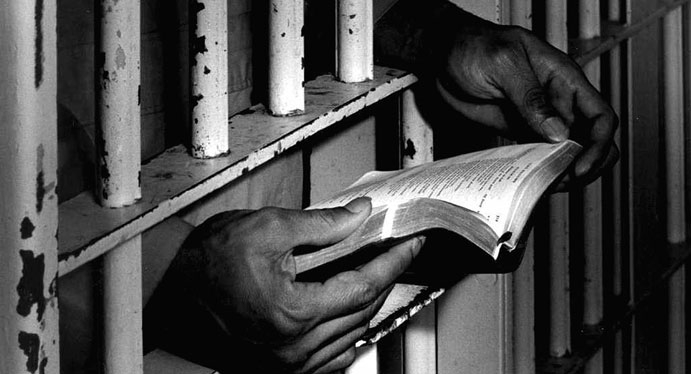 Hands holding a book through the bars of a jail cell.