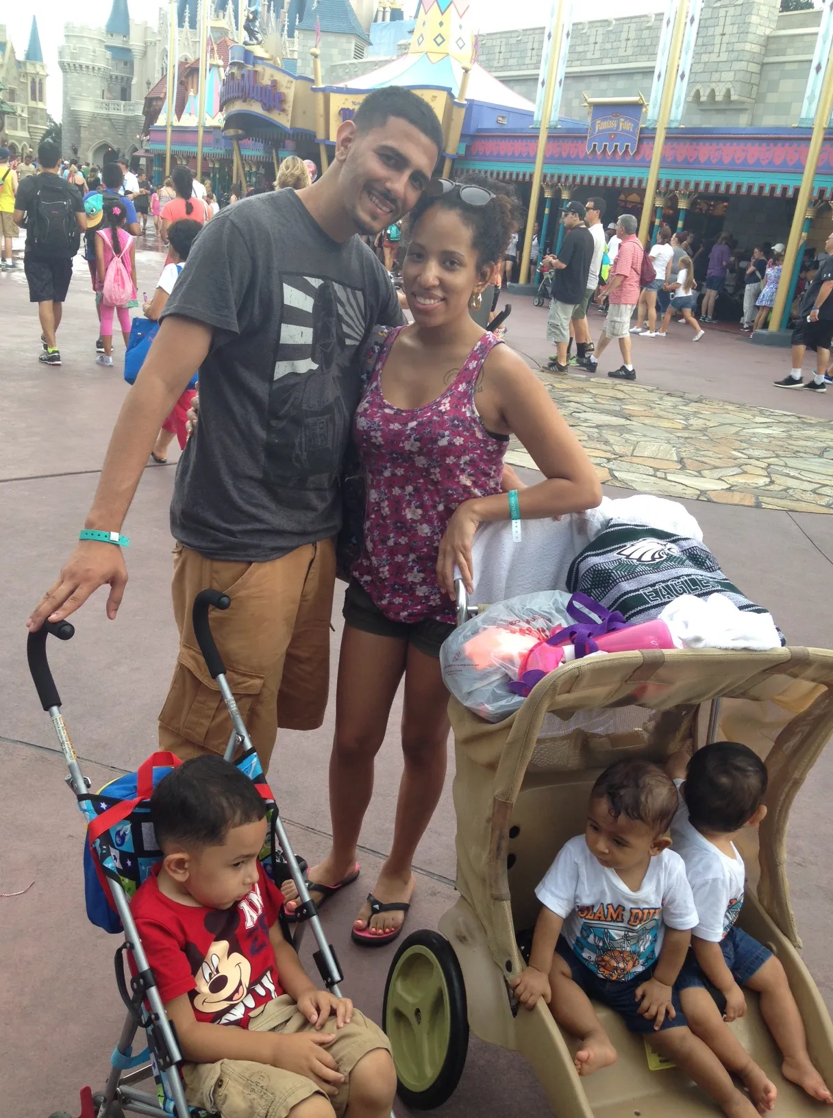 A couple smiling at the camera with two young children in strollers at an amusement park.