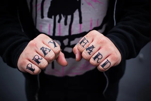 Knuckles tattooed with the words 'pray hard' clenched in front of a hoodie.