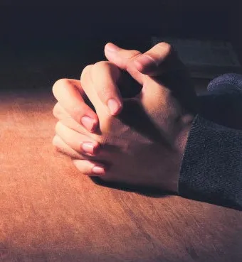 Two hands clasped together on a table illuminated from the side.