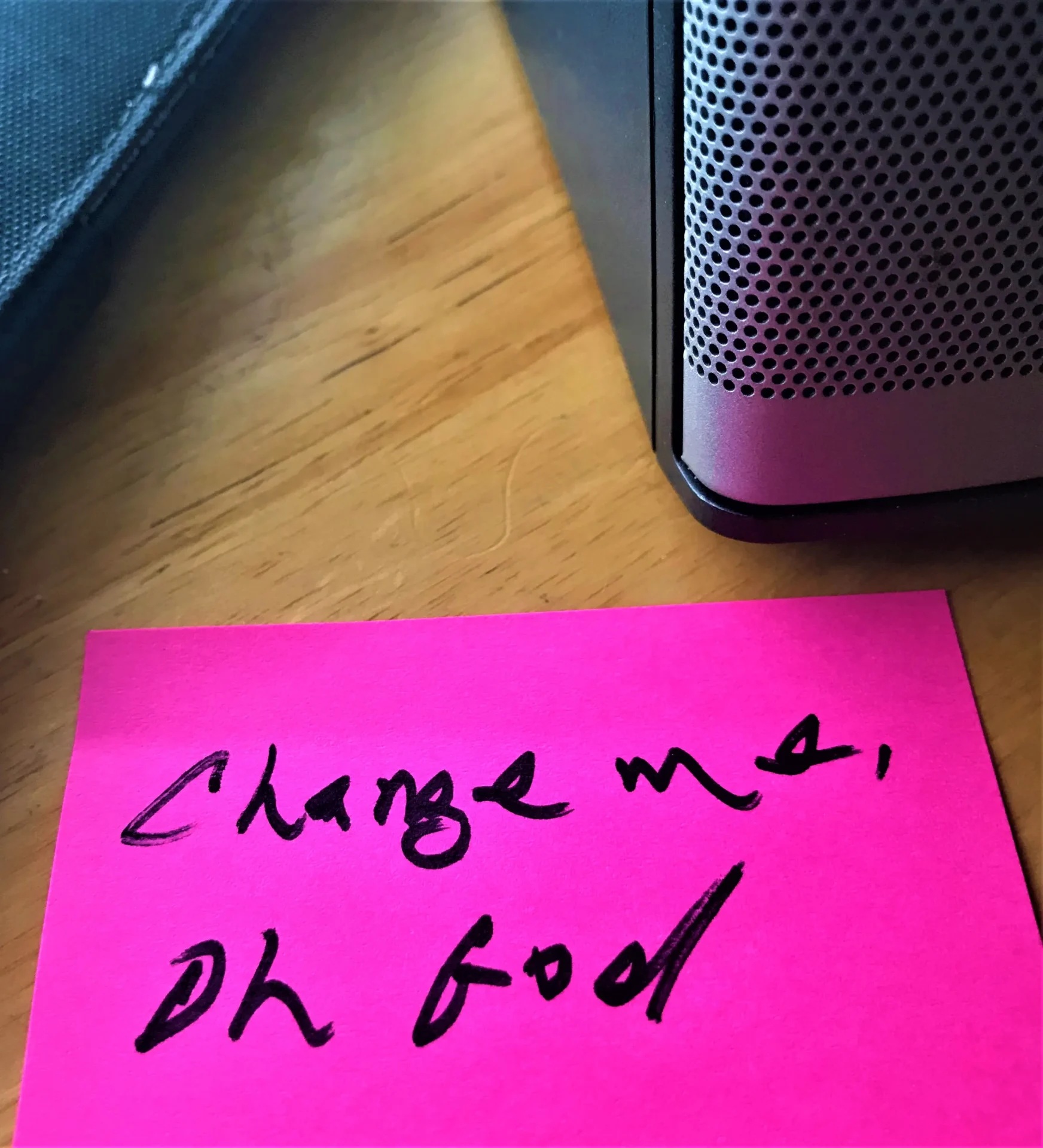 A pink sticky note with the handwritten message "charge me, oh god" placed next to a portable speaker on a wooden table.