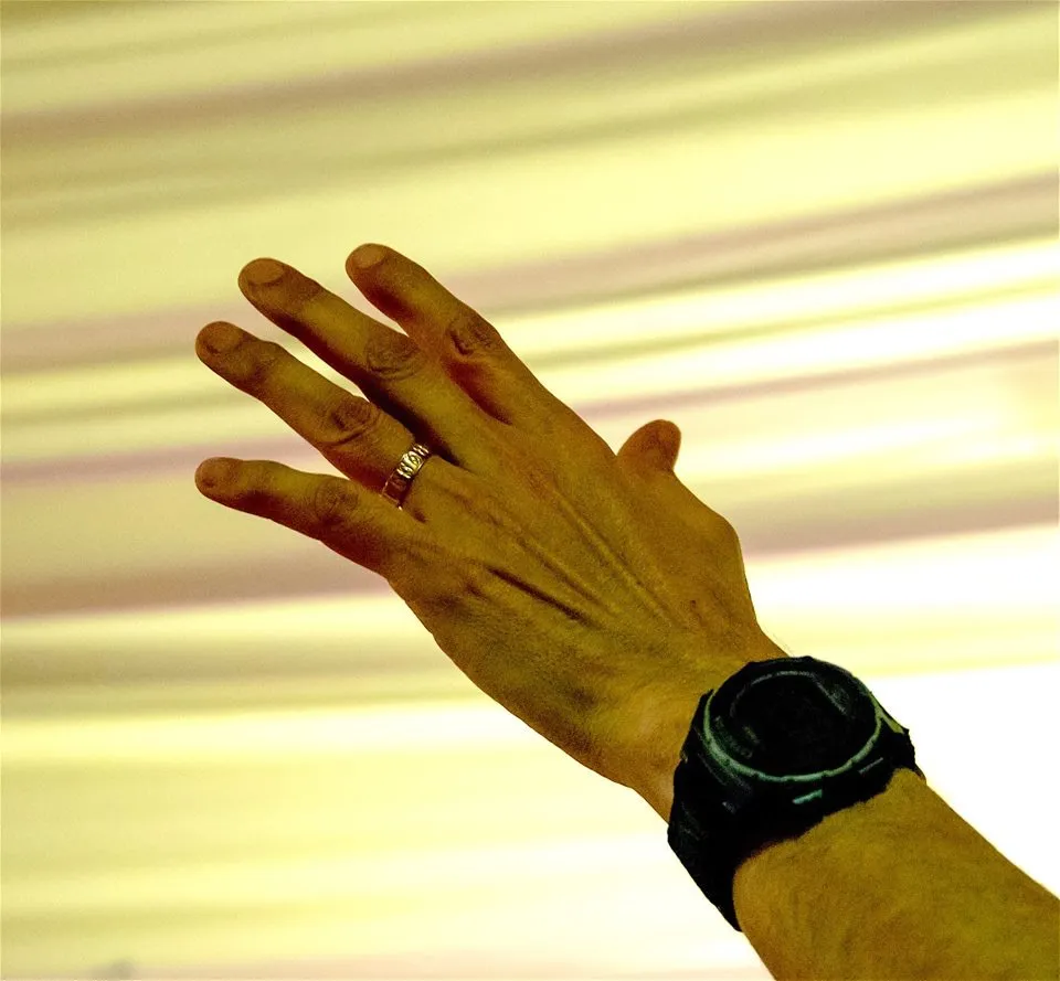 A person's raised hand showing a wristwatch and a ring against a blurred striped background.