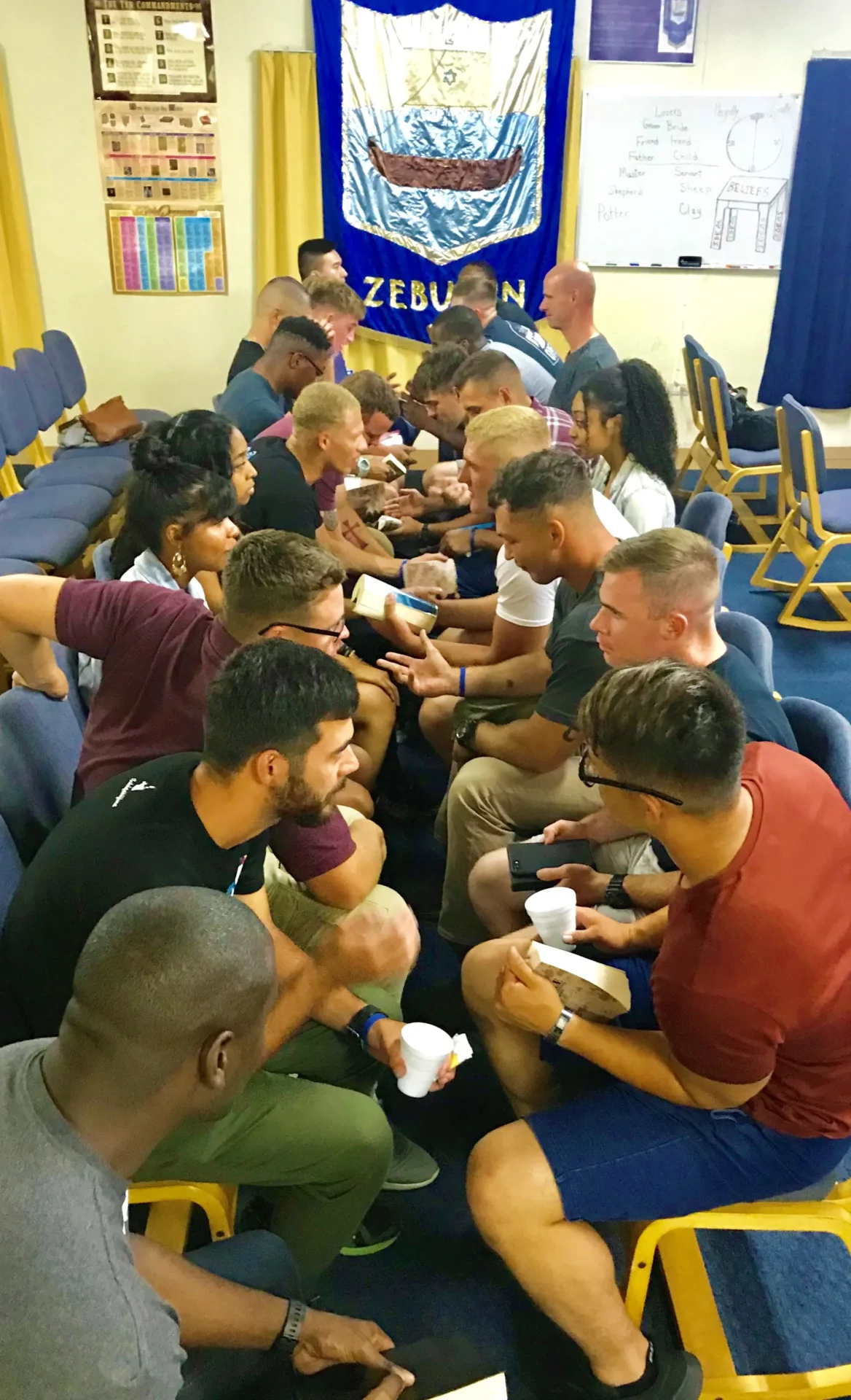 Group of people engaged in a discussion or social activity, seated on chairs arranged in rows inside a room with a blue banner on the wall.