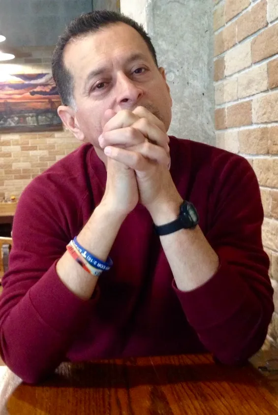 Man sitting at a table with hands clasped, appearing contemplative.