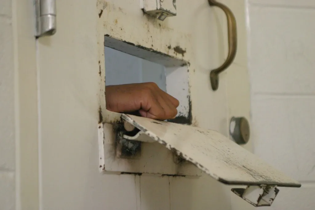 A person's hand is visible through the food tray slot of a weathered prison cell door.