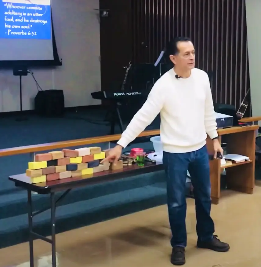 Man giving a presentation with blocks and props on a table in front of a projection screen displaying a bible verse.