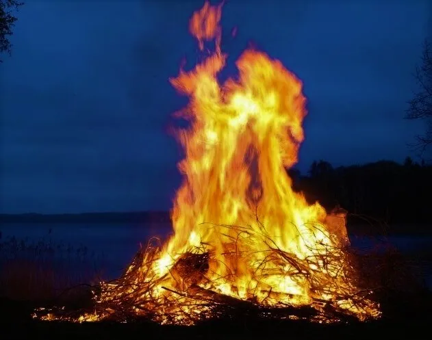 A large bonfire burns brightly against the evening sky by a lakeside.