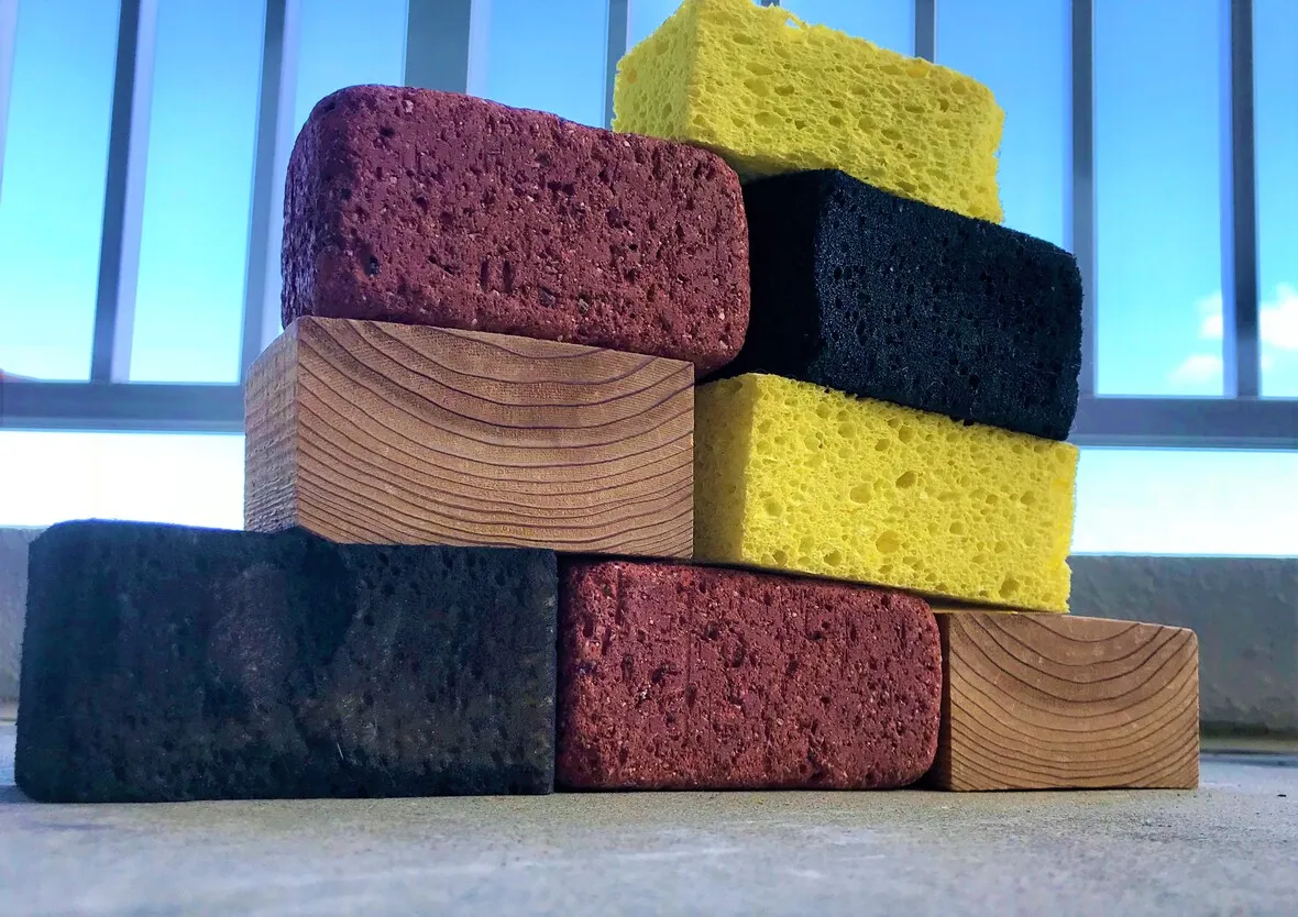 Stack of various textured sponges and wooden blocks arranged in a staggered, tower-like structure on a concrete surface with metal bars in the background.