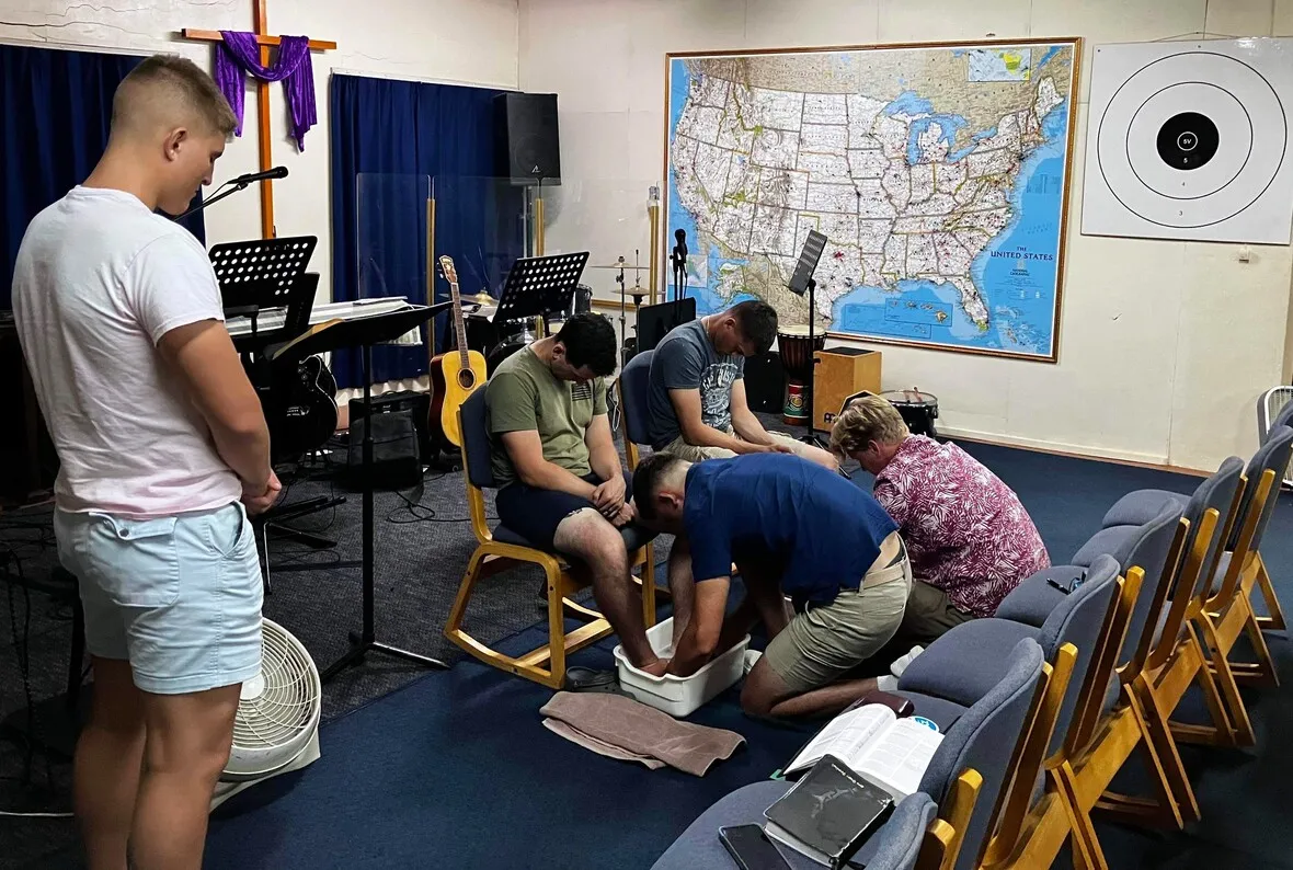 Group of people participating in a foot washing ceremony inside a room with a map and musical equipment.