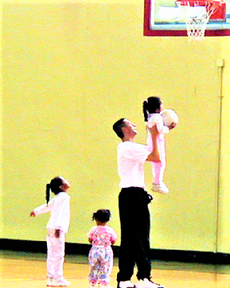 An adult lifting a child toward a basketball hoop as two other children watch.