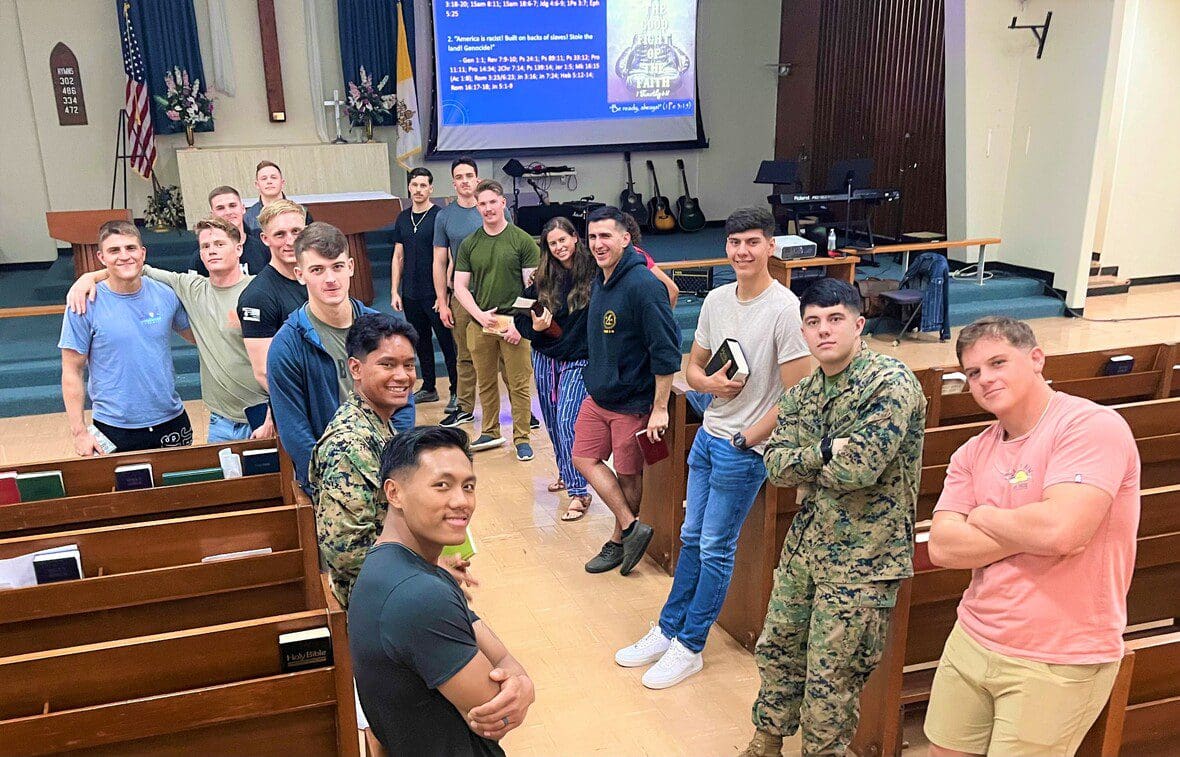 A group of individuals, some in military uniforms, posing for a photograph in a room with rows of benches and a projector screen displaying text.