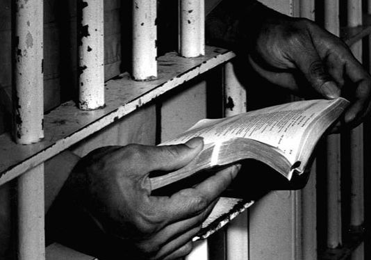 Hands holding a book through the bars of a jail cell.