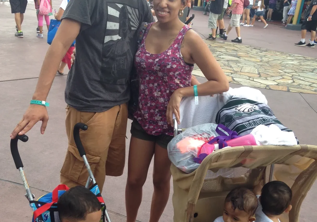 A couple smiling at the camera with two young children in strollers at an amusement park.