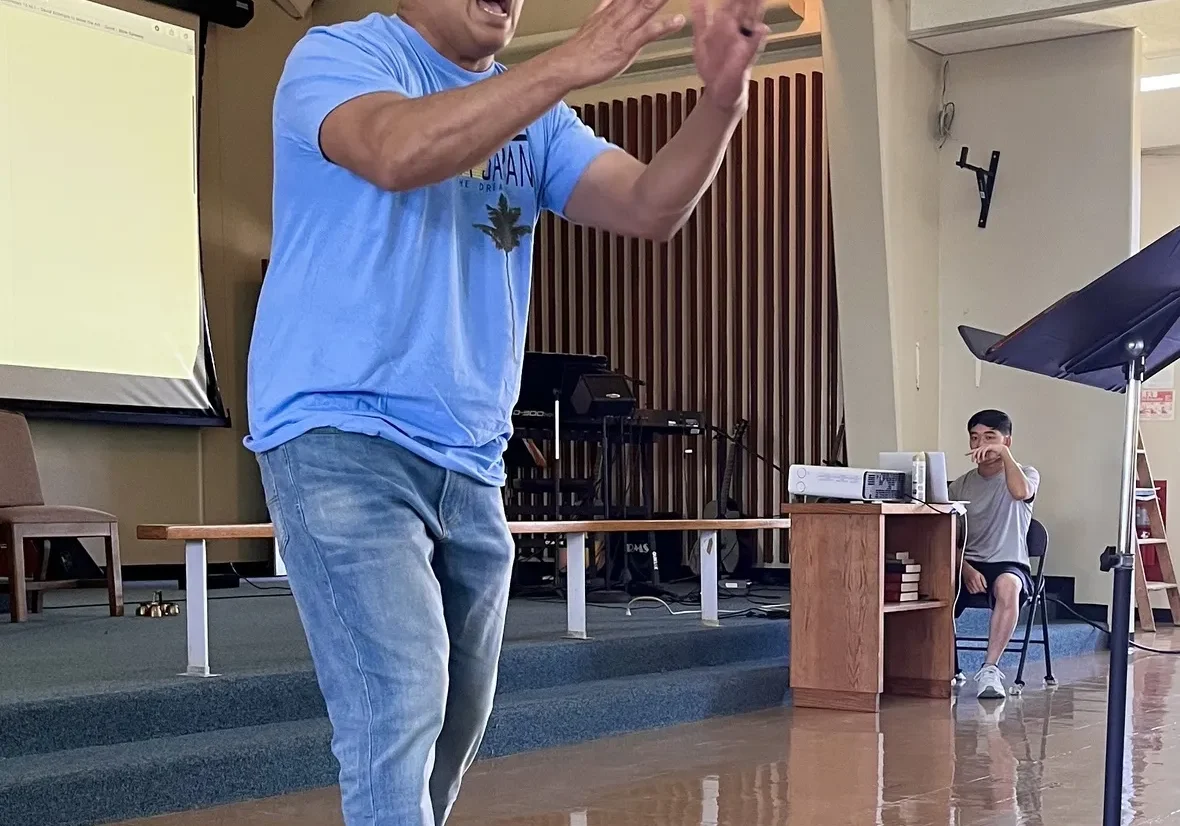 A man enthusiastically speaking and gesturing with his hands in a room with an audience member seated in the background.