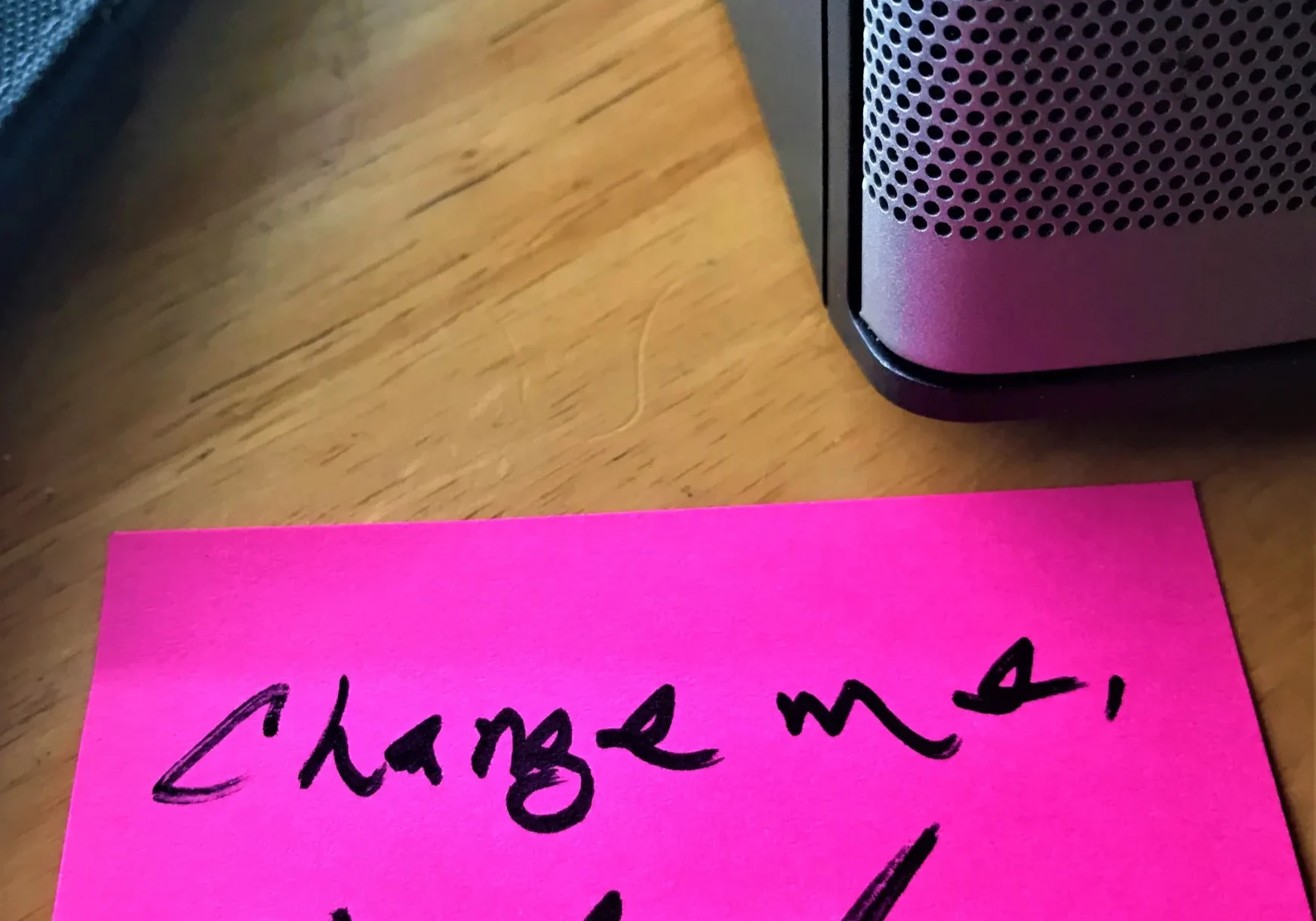 A pink sticky note with the handwritten message "charge me, oh god" placed next to a portable speaker on a wooden table.