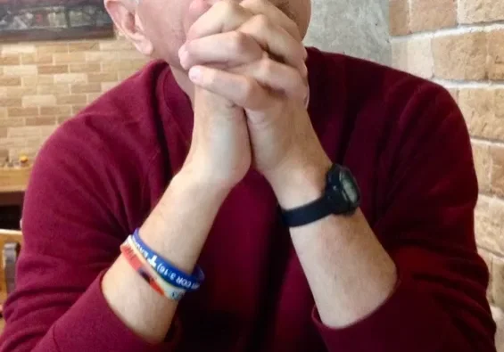 Man sitting at a table with hands clasped, appearing contemplative.