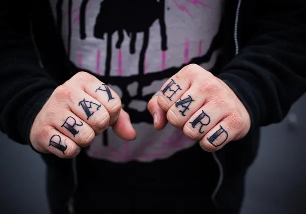 Knuckles tattooed with the words 'pray hard' clenched in front of a hoodie.