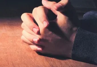 Two hands clasped together on a table illuminated from the side.