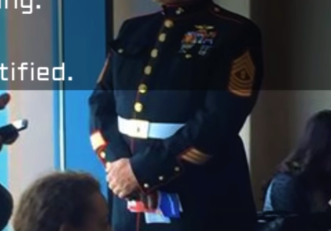 U.s. marine in dress uniform standing in a room with seated guests.