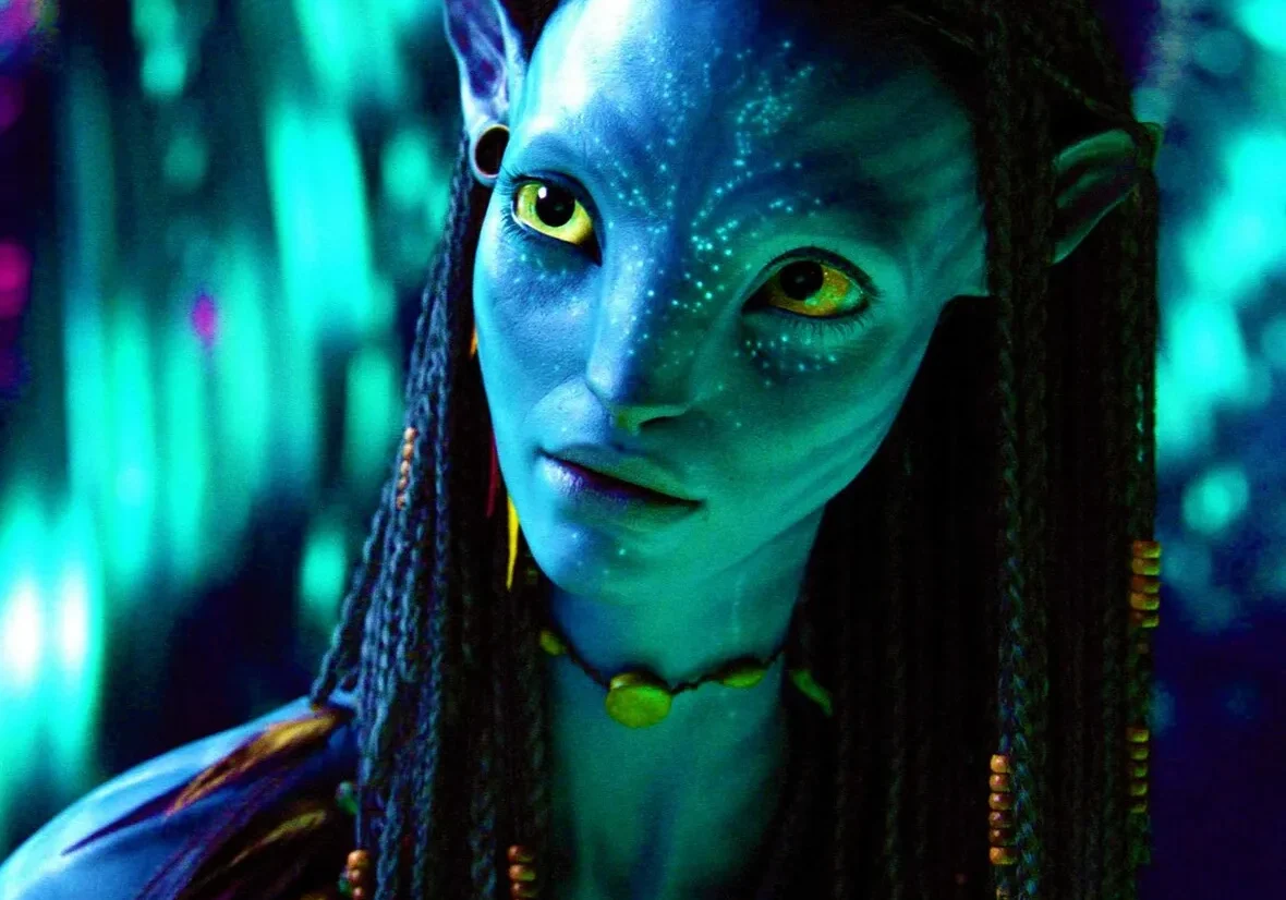 A close-up image of a blue-skinned, wide-eyed, alien character with intricate facial markings and braided hair from a science fiction setting.