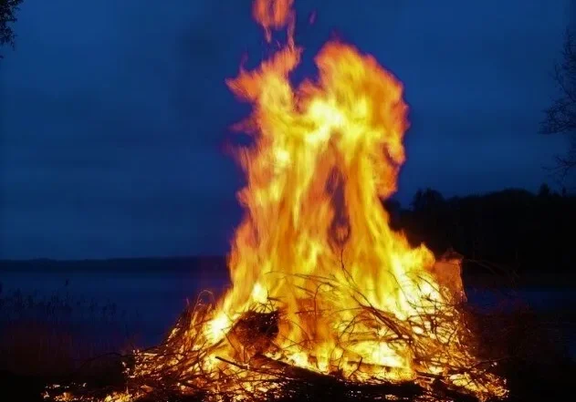 A large bonfire burns brightly against the evening sky by a lakeside.