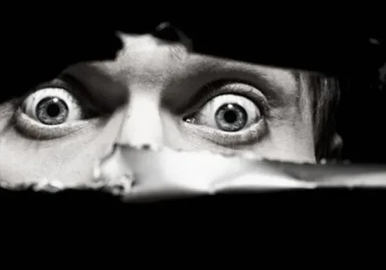 Person peeking through a tear in black material with wide eyes.