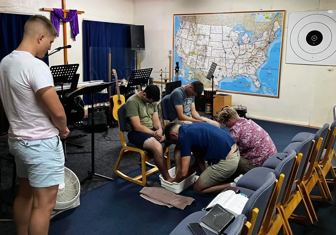 Group of people participating in a foot washing ceremony inside a room with a map and musical equipment.