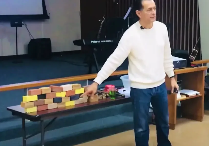 Man giving a presentation with blocks and props on a table in front of a projection screen displaying a bible verse.